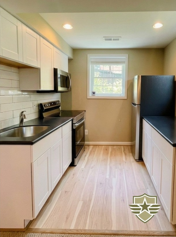 A clean second kitchen in a basement with white cabinets, black countertops, stainless steel appliances, and light wood flooring